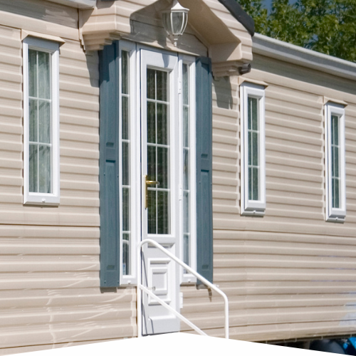 A beige mobile home features a white front door with glass panels and blue shutters. A small white staircase leads up to the door. The scene is bright and sunny.